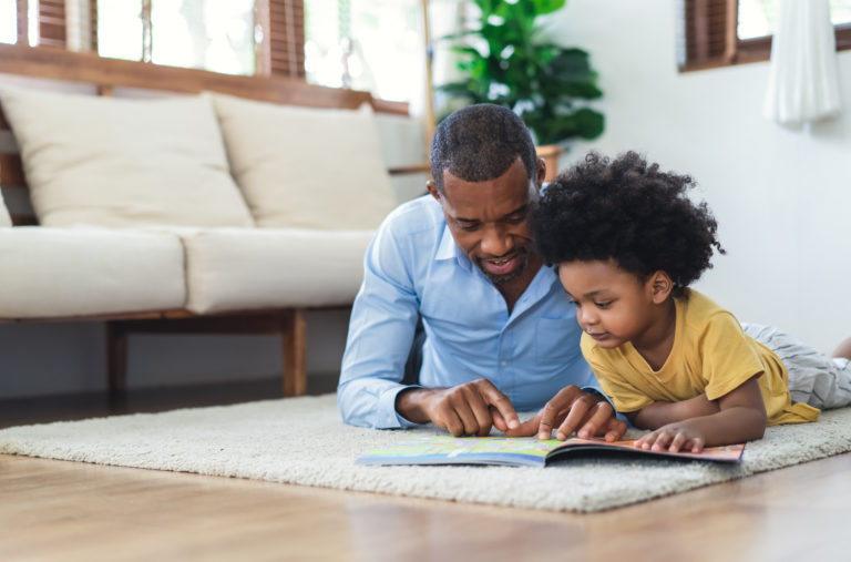 Massachusetts FMLA law concept.Happy African American father and son are reading a book and smiling while lying on floor spending time together at home. Children education and development concept.