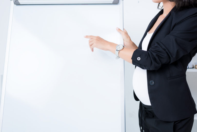 Pregnant employee in black business suit and whit shirt standing next to and pointing to a dry erase board.