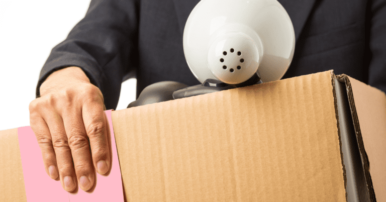 Image of a person wearing a black suit holding a cardboard box with a white desk lamp showing from the top of the box and a "pink slip" in their hand.